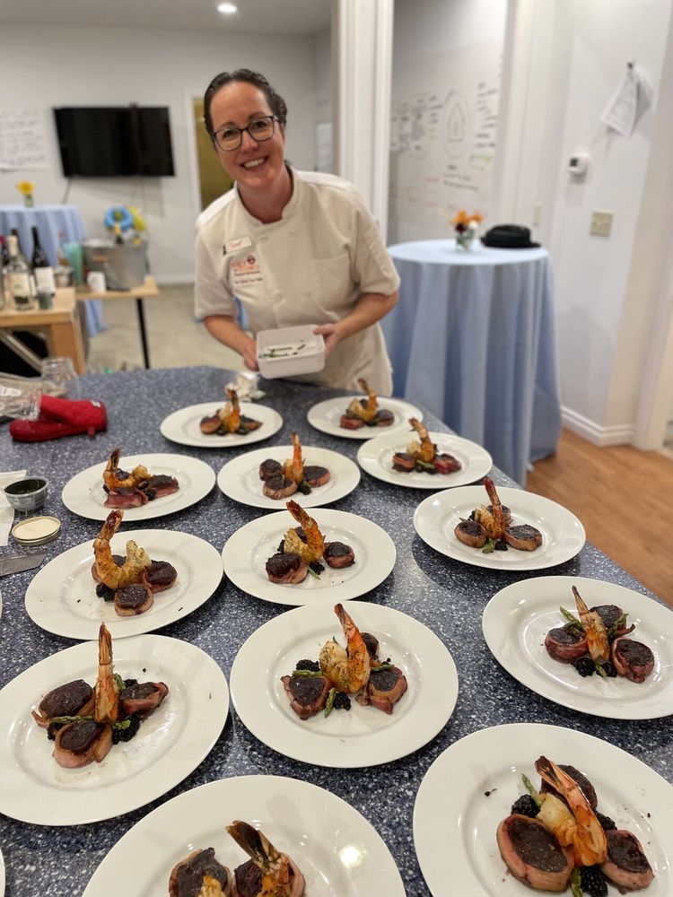 A chef in a white coat stands behind a counter displaying multiple plates of a gourmet shrimp and vegetable dish. The setting appears to be a catered event or cooking class, showcasing exceptional hospitality etiquette.