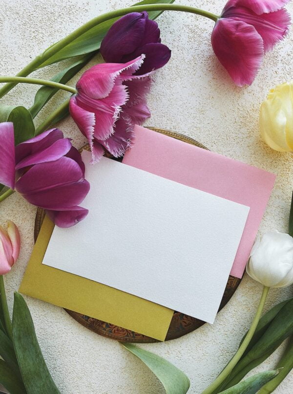 A blank white card placed on top of pink and yellow envelopes, surrounded by various colored tulips on a textured white surface, creates an inviting scene perfect for home entertaining.
