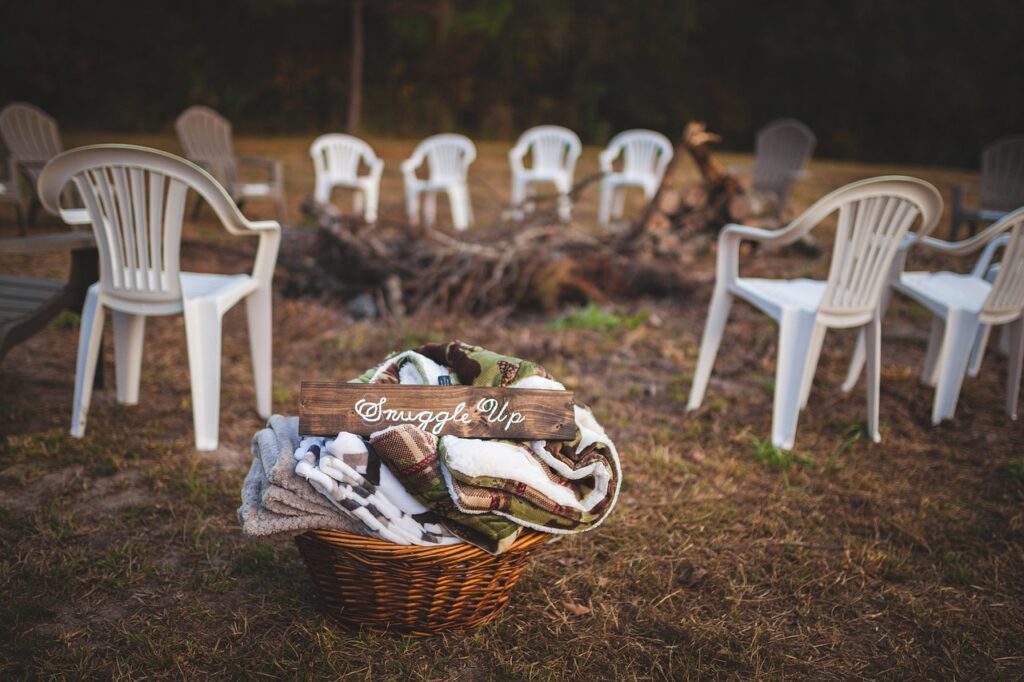 A basket filled with blankets labeled "Snuggle Up" sits in front of a circular arrangement of empty white plastic chairs around a fire pit in a grassy outdoor area.