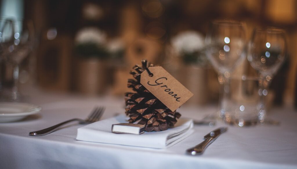 A place setting at a wedding with a folded napkin, a small box, utensils, and a pine cone with a "Groom" tag.