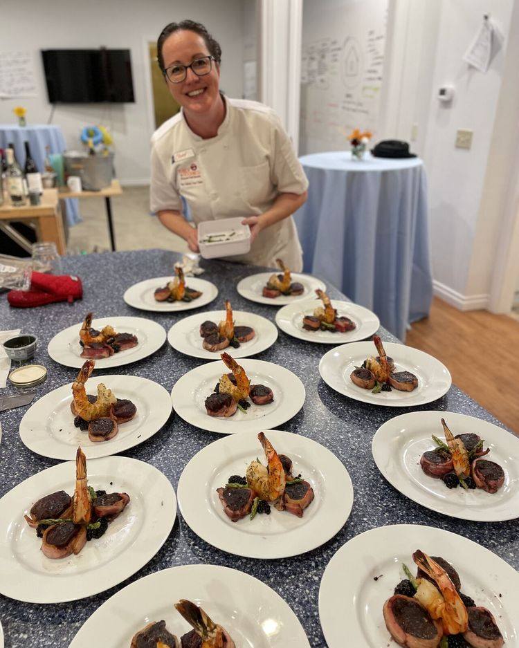 A chef stands behind a counter displaying multiple plates of a gourmet dish, smiling and holding another plate. The dishes appear to feature prawns and vegetables artistically arranged.