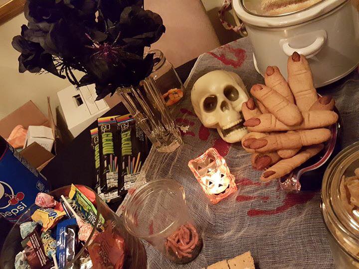A table with a spooky Halloween theme, perfect for a Halloween dinner party, featuring a skull, witch finger cookies, various snacks and candies, a candle, and a crockpot. Black flowers and spooky decorations are spread across the table to set the mood for some eerie party games.
