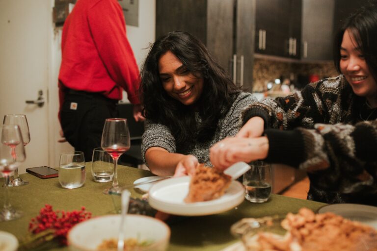 Two women smiling at a dining table, with one serving food to the other, capturing the warmth of Friendsgiving. A red-shirted person stands in the background. Various dishes, glasses, and utensils are spread out on the table.