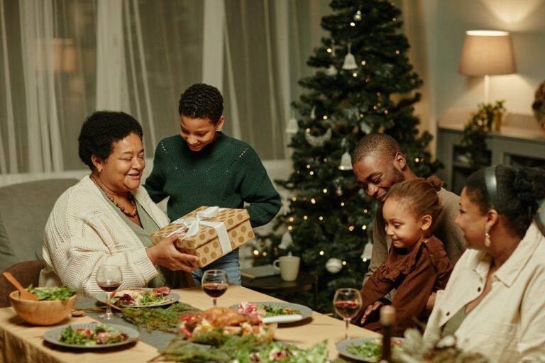 A group of people gather around a festive table with food, as an older woman seated at the head of the table receives a gift from a young boy. A decorated Christmas tree is visible in the background, highlighting the joys of hosting for the holidays and stress-free entertaining.