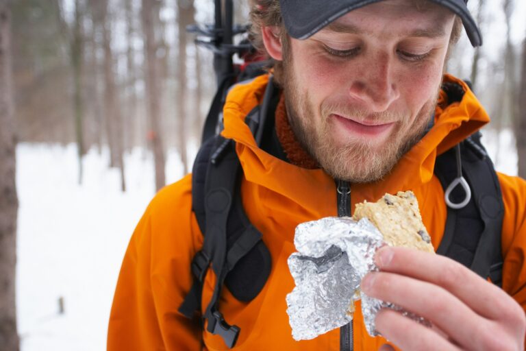 A man wearing a backpack and orange jacket stands in a snowy forest, smiling as he holds and looks at a foil-wrapped sandwich.